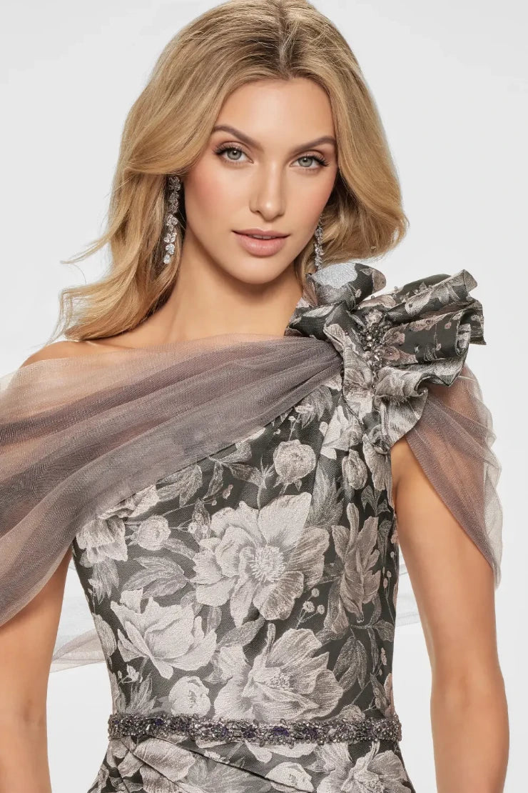Turn heads in this striking trumpet. Its fitted bodice accentuates the waist, while the flared skirt gracefully cascades from the knees, creating that classic trumpet silhouette known for its flattering effect. The dramatic floral brocade adds depth and dimension, while the assymmetrical shoulder design with dreamy tulle and ruffle detail adds an extra touch of glamour. Remove the matching wrap for a sleek and sophisticated look that allows the intricate details of the gown to truly shine.