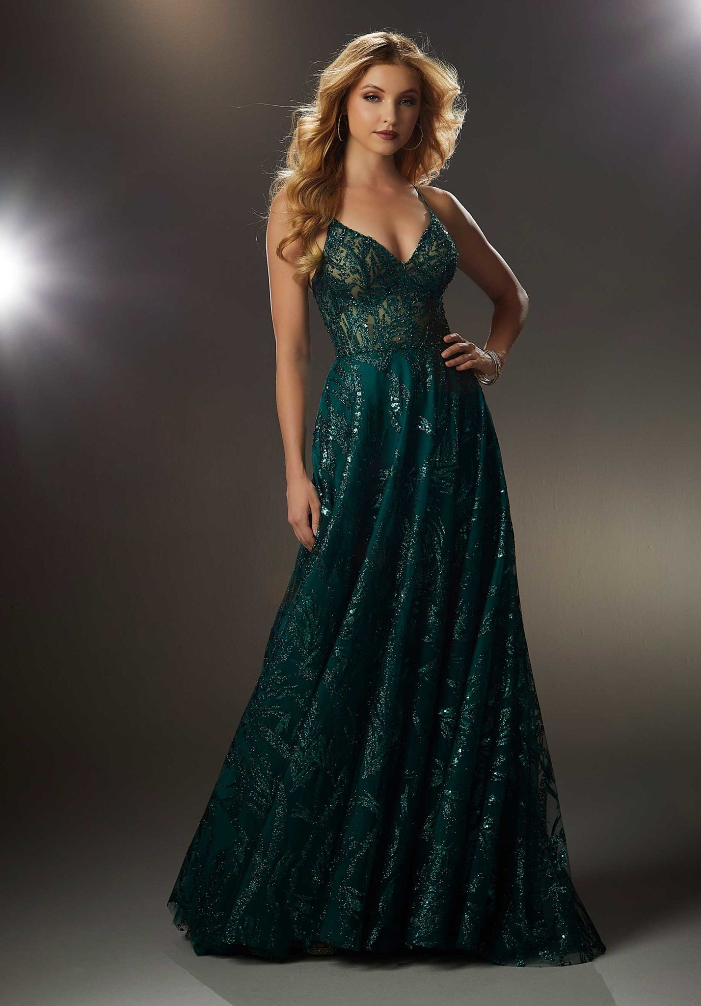 Classic A-line prom dress features a sheer deep-v bodice and flowing skirt designed with sparkling patterned glitter and blingy beaded trim. The spaghetti straps cross in the back meeting up with the lace-up corset back.